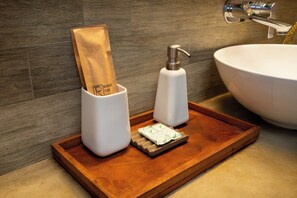 Un lavamanos de baño con un lavabo blanco, una bandeja de madera con dispensador de jabón y una pastilla de jabón, y un porta toallas de papel con una toalla de papel marrón.
