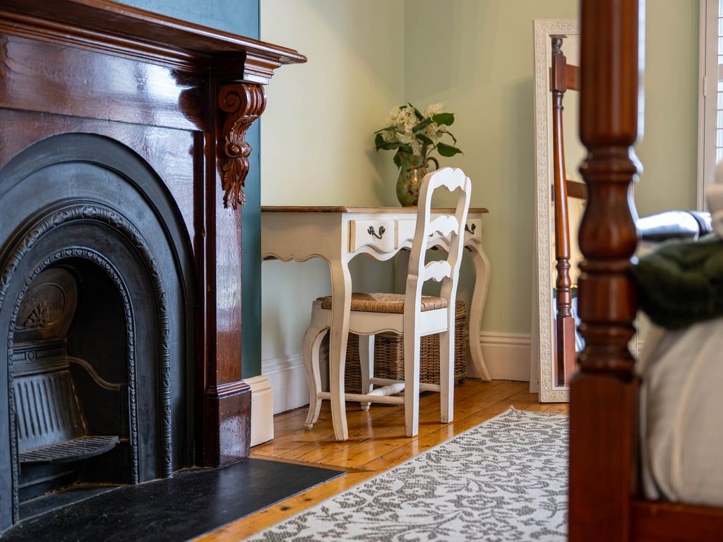 A room with a fireplace, a white chair, a small table with a vase, and a patterned rug.
