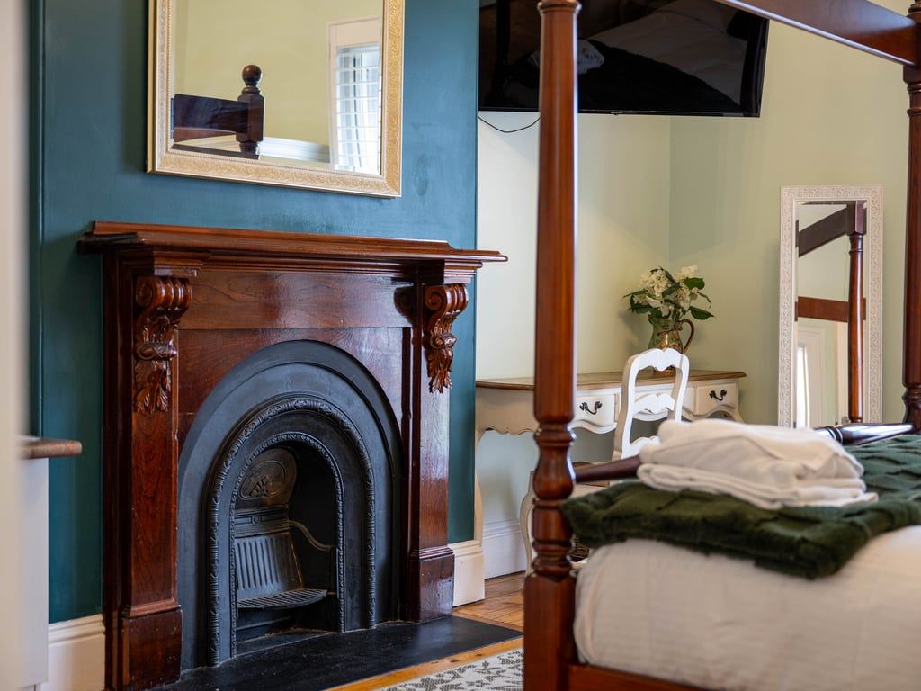 A bedroom with a four-poster bed, a fireplace, a mirror, and a bedside table with a plant.