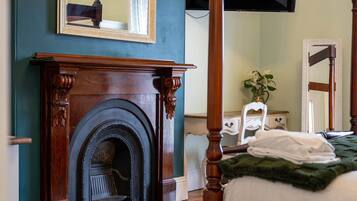 A bedroom with a four-poster bed, a fireplace, a mirror, and a bedside table with a plant.