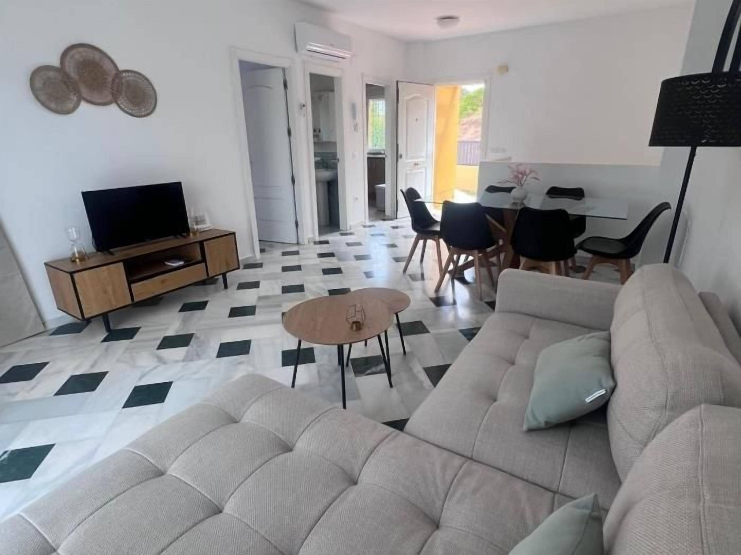 A living room with a sofa, a coffee table, a TV on a wooden cabinet, and a dining area with chairs and a table.