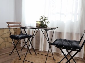 Ein Zimmer mit Tisch und zwei Stühlen, einer Topfpflanze auf dem Tisch und einem Fenster mit durchsichtigen Vorhängen.