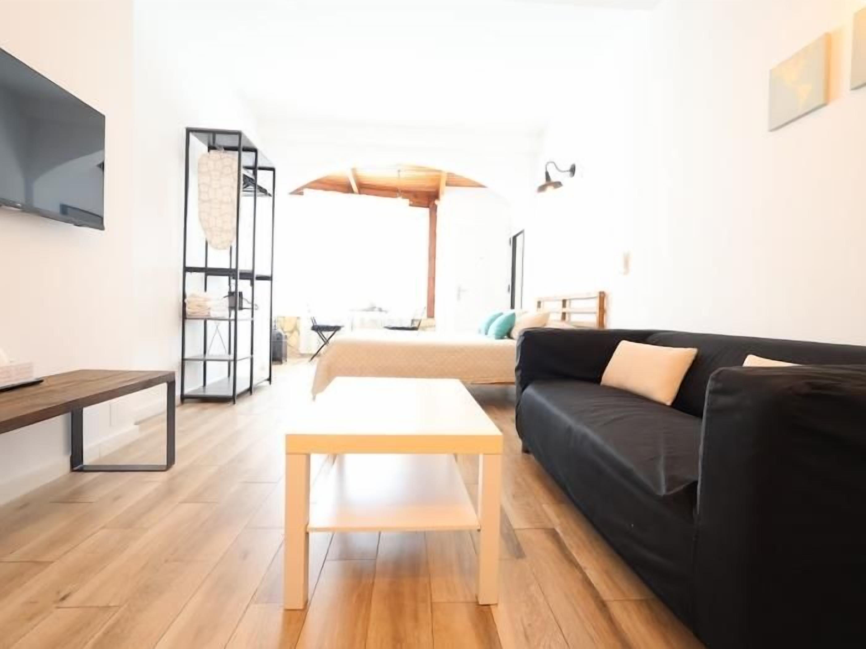 A modern living room with a wooden floor, a black sofa, a wooden coffee table, and a TV mounted on the wall.