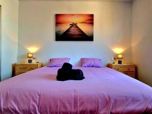 Ein Schlafzimmer mit einem Bett, zwei Nachttischen, einem Gemälde mit einem Sonnenuntergang über Wasser und einem Hund, der auf dem Bett liegt.