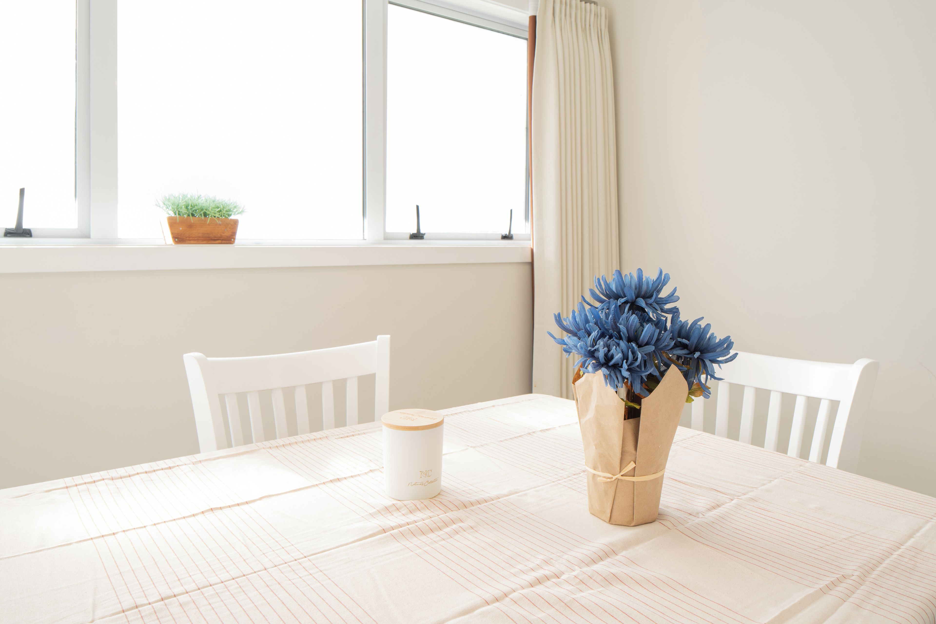 A table with a white tablecloth, a white chair, a vase with blue flowers, and a white candle on the table.