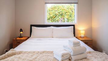 A neatly made bed with white linens and a fluffy beige rug, a window with a view of a tree, and a bedside lamp on a wooden nightstand.