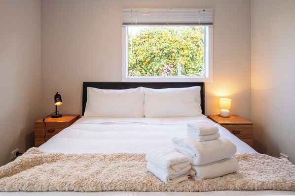 A neatly made bed with white linens and a fluffy beige rug, a window with a view of a tree, and a bedside lamp on a wooden nightstand.