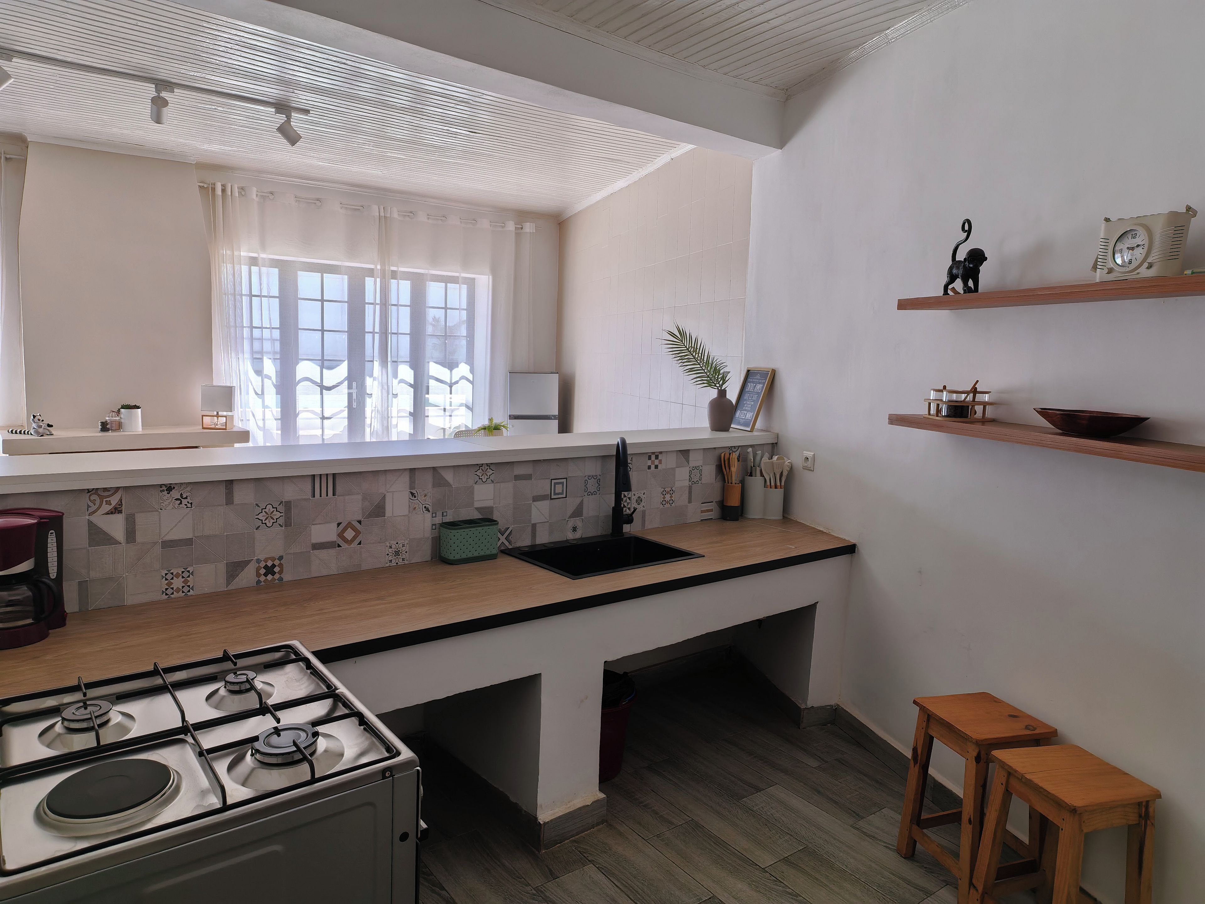 A modern kitchen with a gas stove, a window with sheer curtains, and a shelf with kitchen utensils.