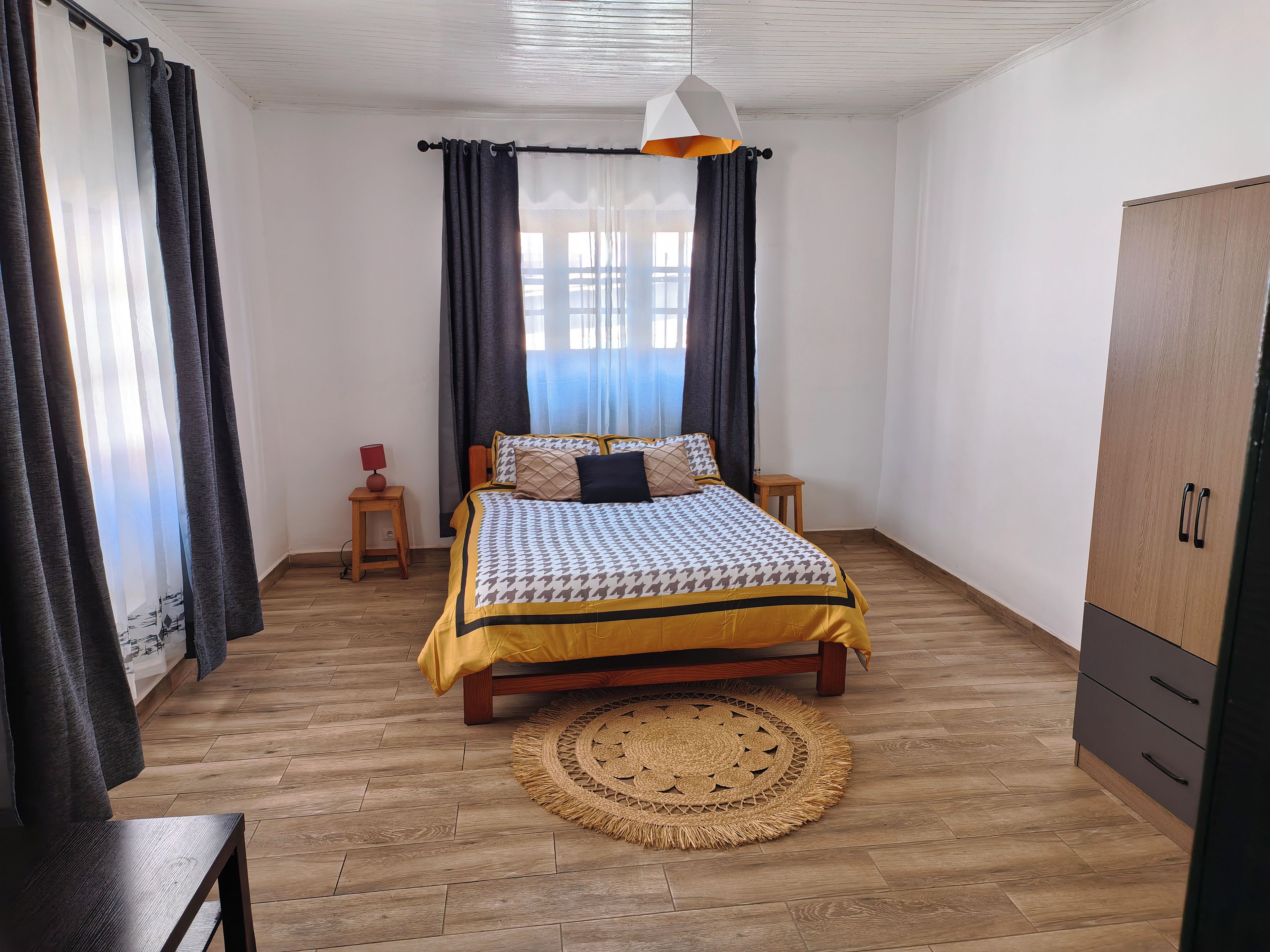 A bedroom with a bed, a wooden wardrobe, a bedside table, a lamp, and a window with curtains.