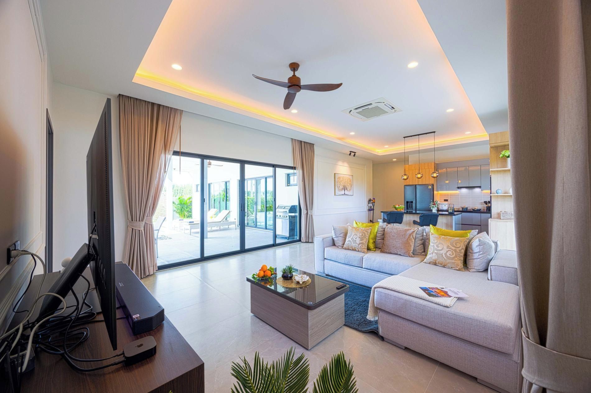A modern living room with a flat-screen TV, a ceiling fan, a glass coffee table, and a sofa with cushions.