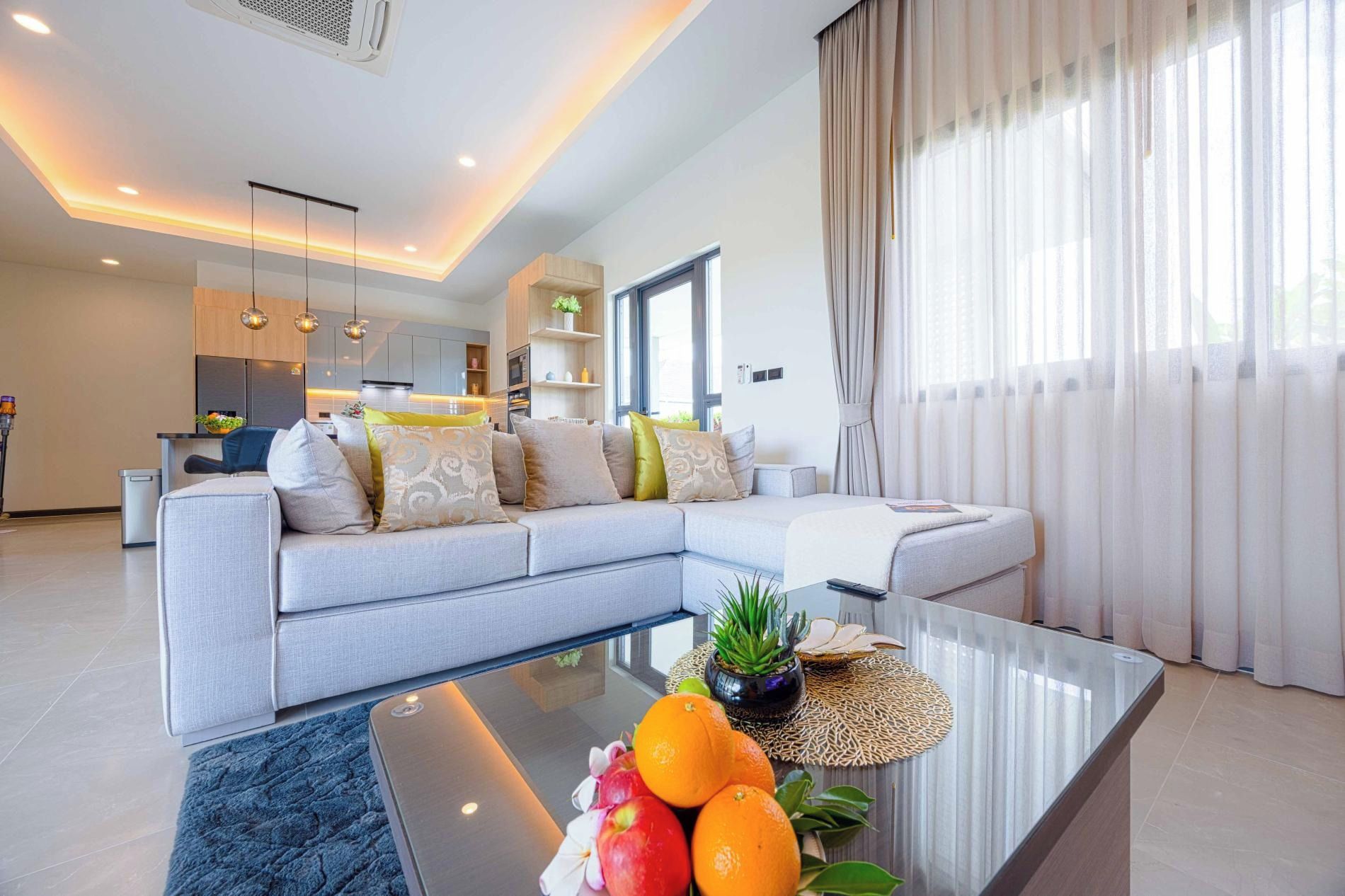 A modern living room with a sectional sofa, a glass coffee table with fruits and a plant, and a kitchen in the background.