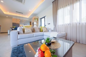 A modern living room with a sectional sofa, a glass coffee table with fruits and a plant, and a kitchen in the background. - Modern 3BR Villa with Private Pool (NR1) (Hua Hin)