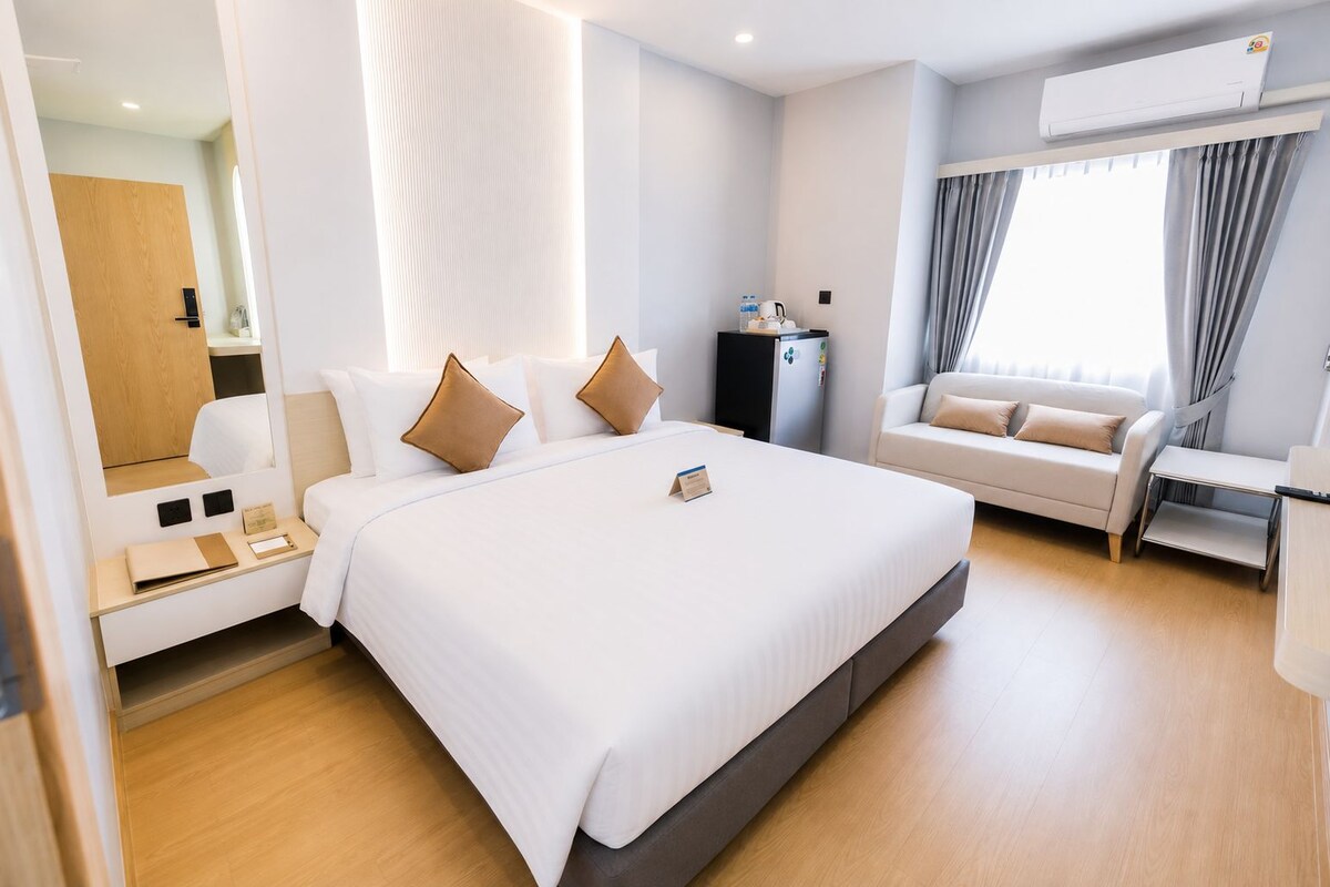 A modern hotel room with a large bed, a sofa, a desk, and an air conditioning unit.