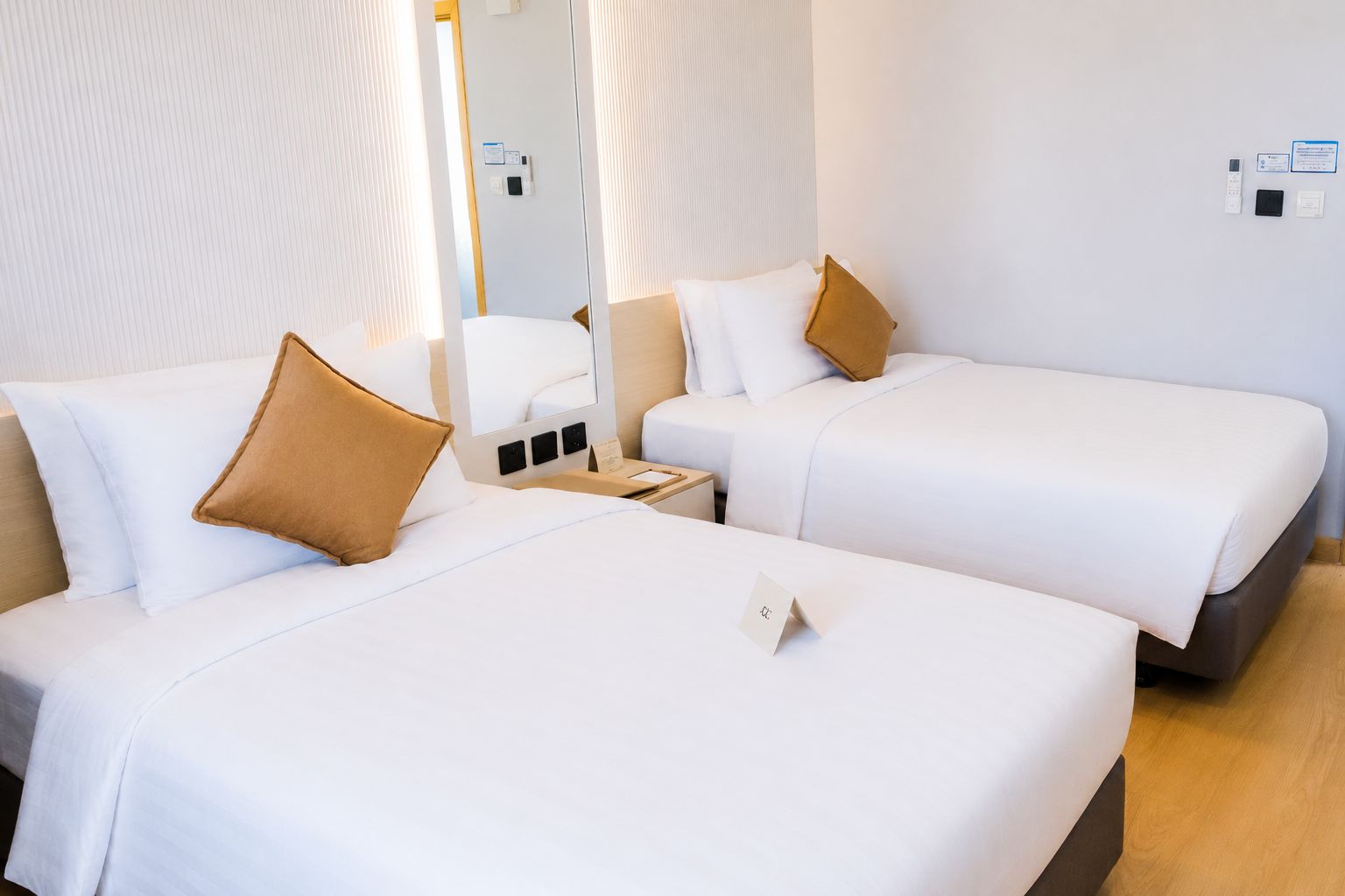 A hotel room with two beds, each with white bedding and a single brown pillow. There is a bedside table with a lamp and a small card.