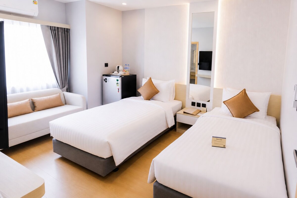 A hotel room with two beds, a desk, a minibar, and a large window with curtains.