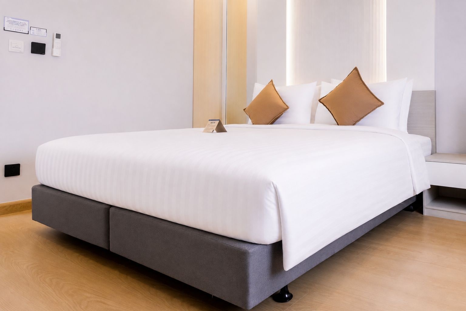 A modern hotel room with a large bed, grey upholstered headboard, white bedding, and a bedside table with a lamp.
