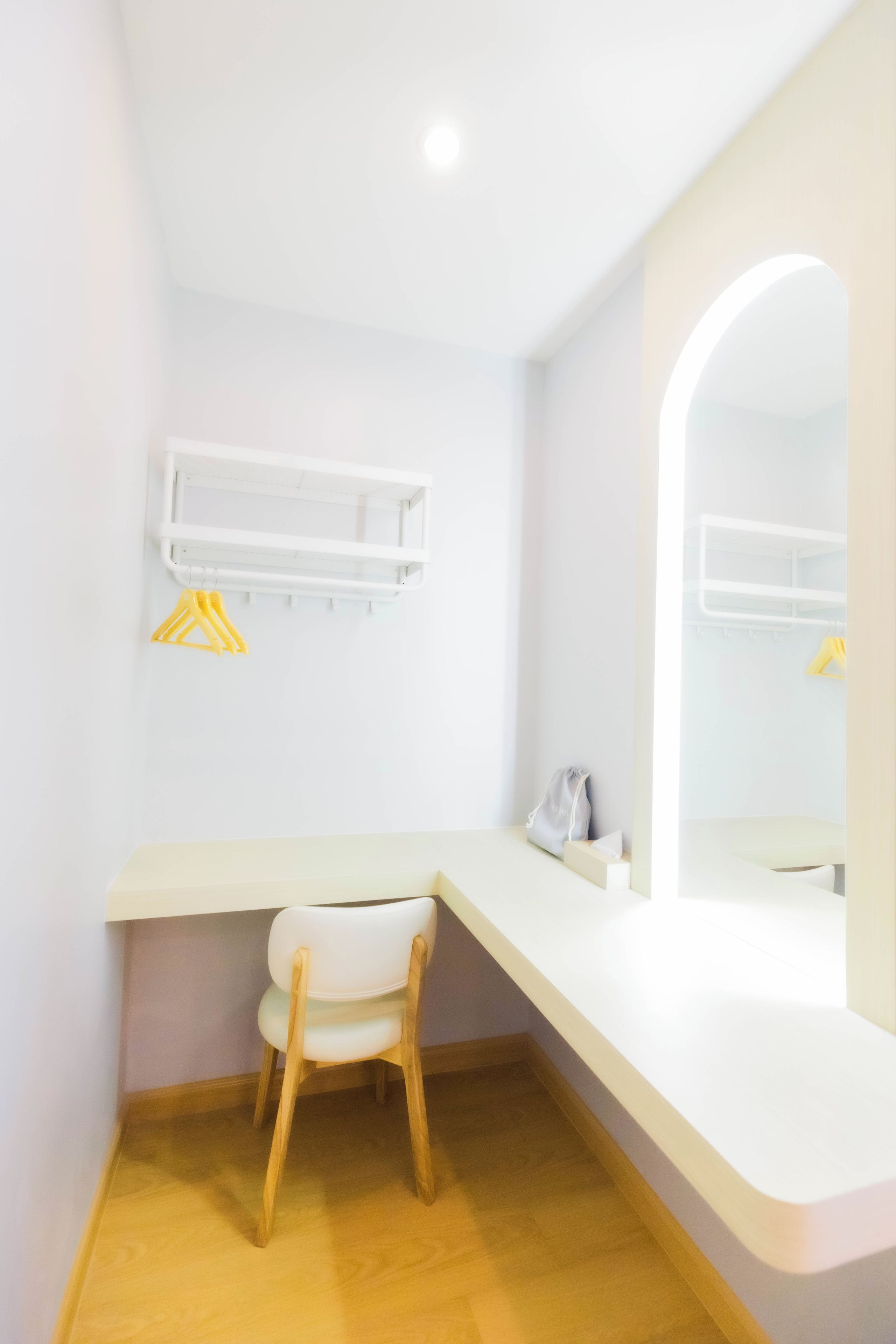 A small, modern bathroom with a white vanity, a wooden chair, a mirror, and a shelf with hangers.