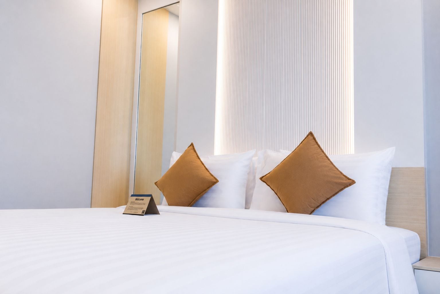 A hotel room with a large bed, two pillows, a nightstand, a mirror, and a window with vertical blinds.