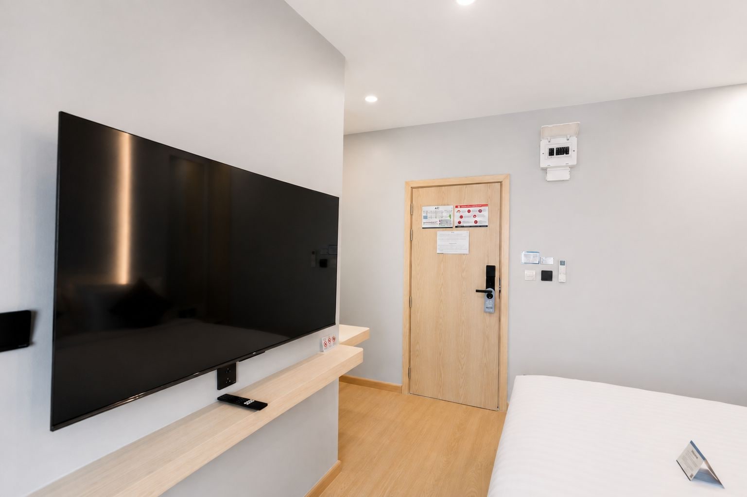 A modern hotel room with a flat-screen TV mounted on the wall, a wooden door with a card holder, and a bedside table with a remote control.