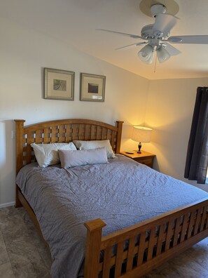 Een slaapkamer met een houten bed, een plafondventilator, twee ingelijste foto’s aan de muur, een bedlamp en een raam met gordijnen.