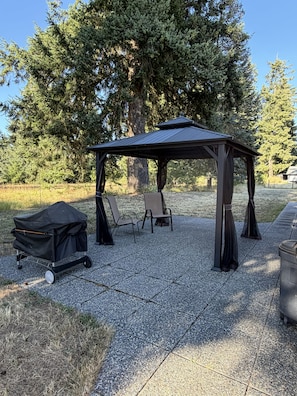 Een prieeltje met dak en vier stoelen, gelegen op een grindpad, met bomen op de achtergrond.