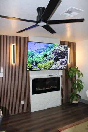 Ein Wohnzimmer mit Flachbildfernseher über einem Kamin, Deckenventilator und Zimmerpflanze.