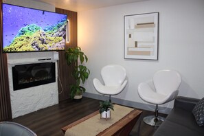 Ein Wohnzimmer mit Flachbildfernseher, zwei weißen Sesseln, einem kleinen Tisch mit Pflanze und einem gerahmten abstrakten Kunstwerk an der Wand.