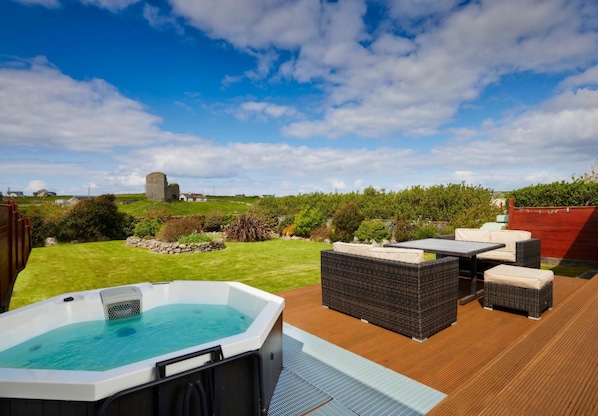 Ein Whirlpool auf einer Holzdielen-Terrasse mit Gartenmöbeln, umgeben von einer Rasenfläche und einem rotem Zaun.