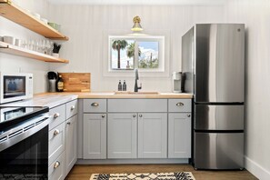 A modern kitchen with a stainless steel refrigerator, a microwave, and a stove. There is a window above the sink, and a wooden shelf with glasses and a plant.