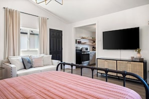A modern bedroom with a bed, a sofa, a TV, and a kitchenette in the background.