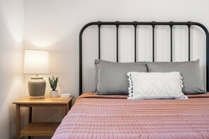 A neatly made bed with a pink quilt, a bedside lamp, a small potted plant, and a white decorative pillow.