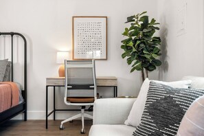 A modern bedroom with a bed, a desk, a chair, a potted plant, and a wall-mounted artwork.