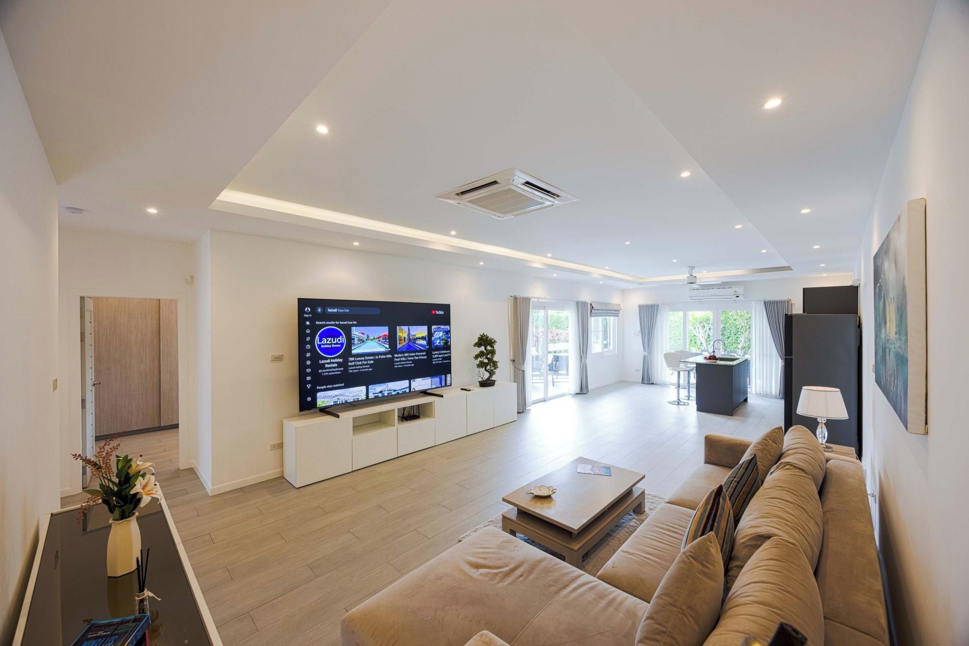 A modern living room with a flat-screen TV, a sofa, a coffee table, and a kitchen area in the background.