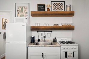 A kitchen with white cabinets, a gas stove, a sink, and a coffee maker. There are wooden shelves above the counter with decorative items and a framed botanical illustration. - Arcadia Studio | Private Entry, Desk + Fast WiFi (Phoenix)