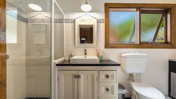 A bathroom with a glass shower enclosure, a sink with a mirror above it, a toilet, and a window with a view of greenery.