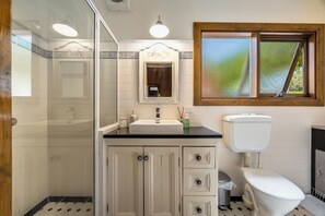 A bathroom with a glass shower enclosure, a sink with a mirror above it, a toilet, and a window with a view of greenery. - Vineyard Charm House | McLaren Vale Retreat (Blewitt Springs)