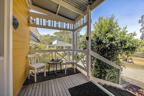 A covered outdoor seating area with a bench and table, a wooden deck, and a view of a building labeled 'RJ Vineyard Cellar Door' in the background. - Vineyard Charm House | McLaren Vale Retreat (Blewitt Springs)