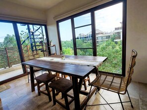 A dining area with a wooden table and chairs, large windows with a view of greenery and buildings, and a staircase with a railing. - Luxury 2BR Penthouse in the Jungle. Nature, Pools & Cenotes in Tulum (Tulum)
