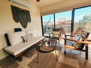 A modern living room with a sofa, chairs, a coffee table, and a large window offering a view of a thatched-roof structure. - Luxury 2BR Penthouse in the Jungle. Nature, Pools & Cenotes in Tulum (Tulum)
