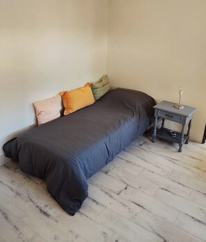 Een enkel bed met een donkere matras en meerdere kussens in een kamer met een betegelde vloer en een klein nachtkastje met een lamp.