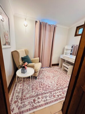 A living room with a beige armchair, a small round table with a vase, a patterned rug, and a white desk with shelves.