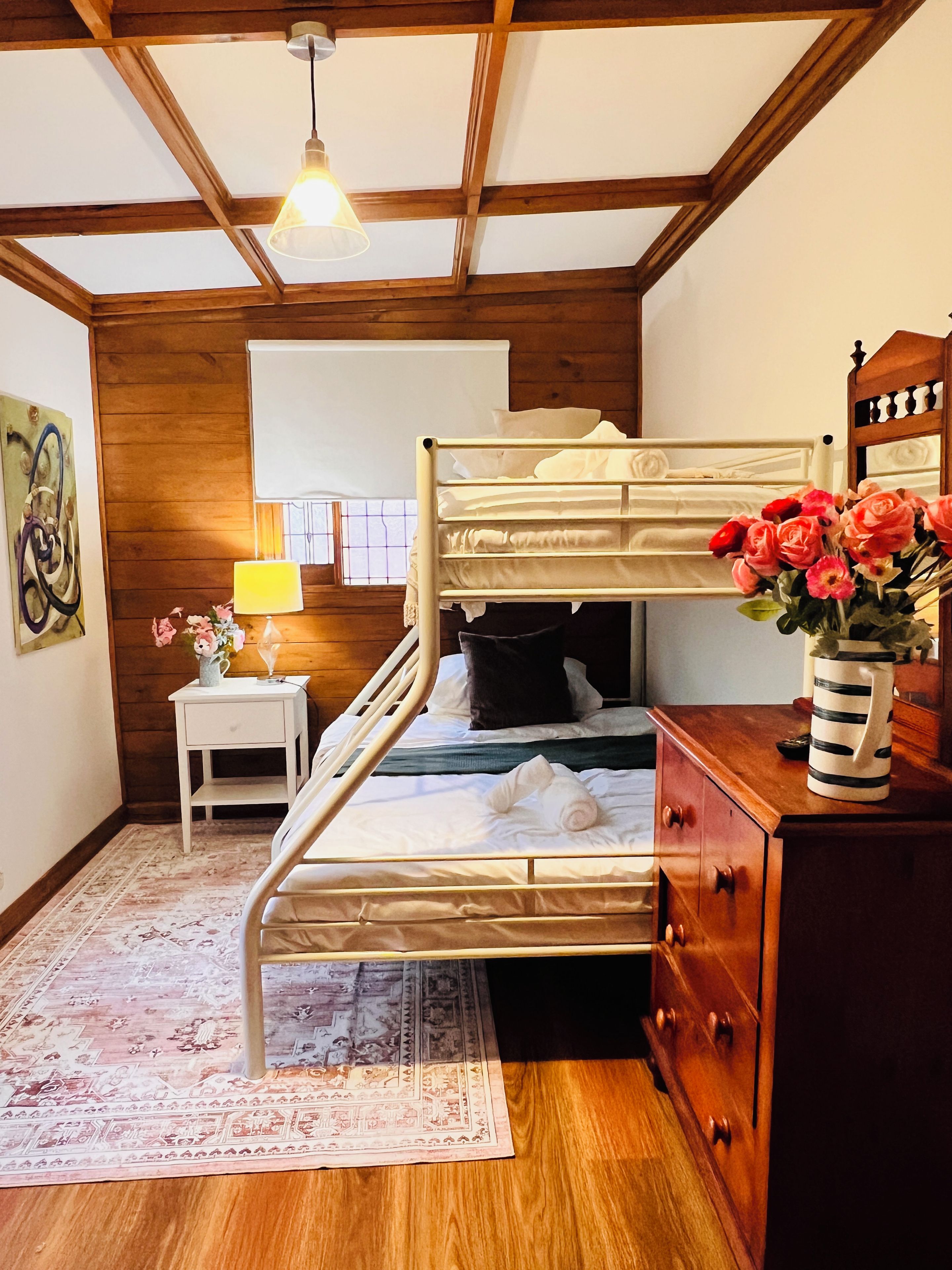 A bunk bed room with a wooden ceiling, a bedside table with a lamp, a dresser, and a rug.