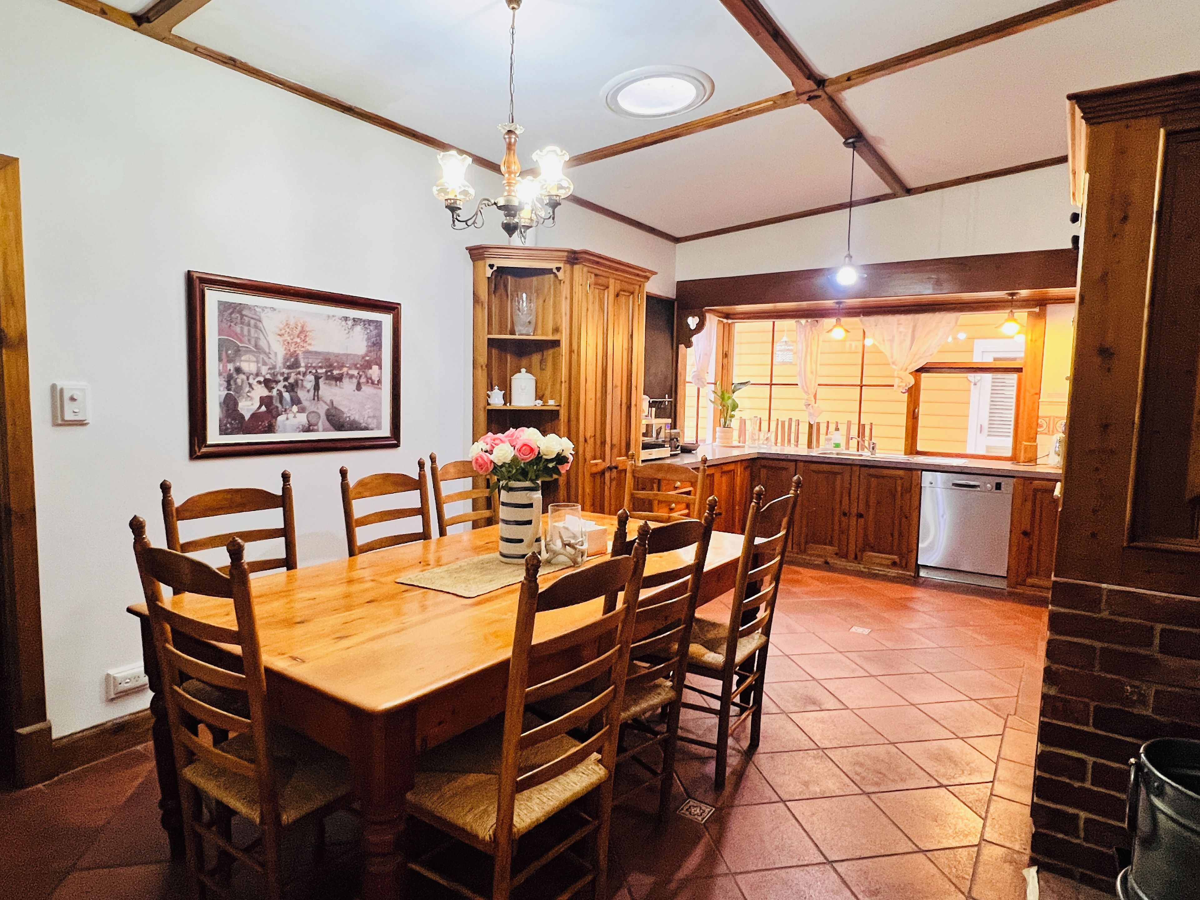 A dining area with a wooden table, chairs, a chandelier, a framed picture, and a kitchen in the background.