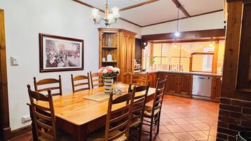 A dining area with a wooden table, chairs, a chandelier, a framed picture, and a kitchen in the background.