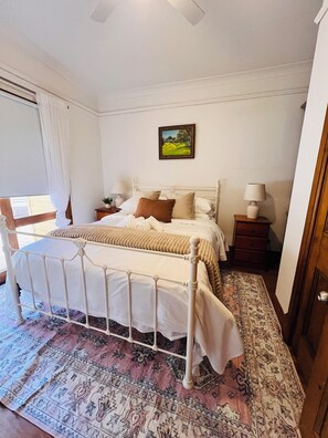 Une chambre à coucher avec un lit, une table de chevet, une lampe, une fenêtre avec des rideaux et un tableau au mur.