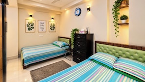 A bedroom with two single beds, a dresser, a clock, wall-mounted lights, and a potted plant. - AST Guest House (Dhaka)