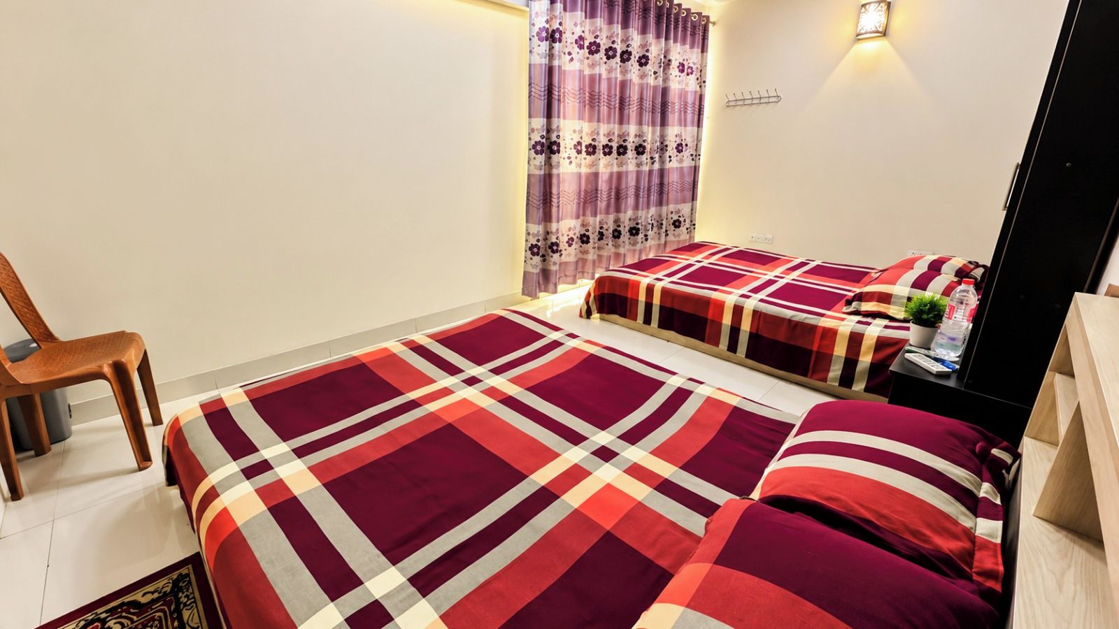 Un dormitorio con dos camas, una silla, una mesita de noche con una planta y una lámpara fijada a la pared.