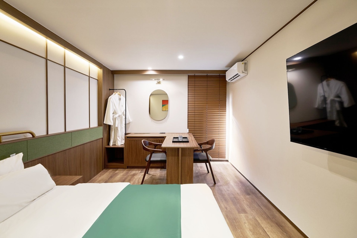 Habitación de hotel con cama, escritorio, silla, perchero para bata y ventana con persianas.