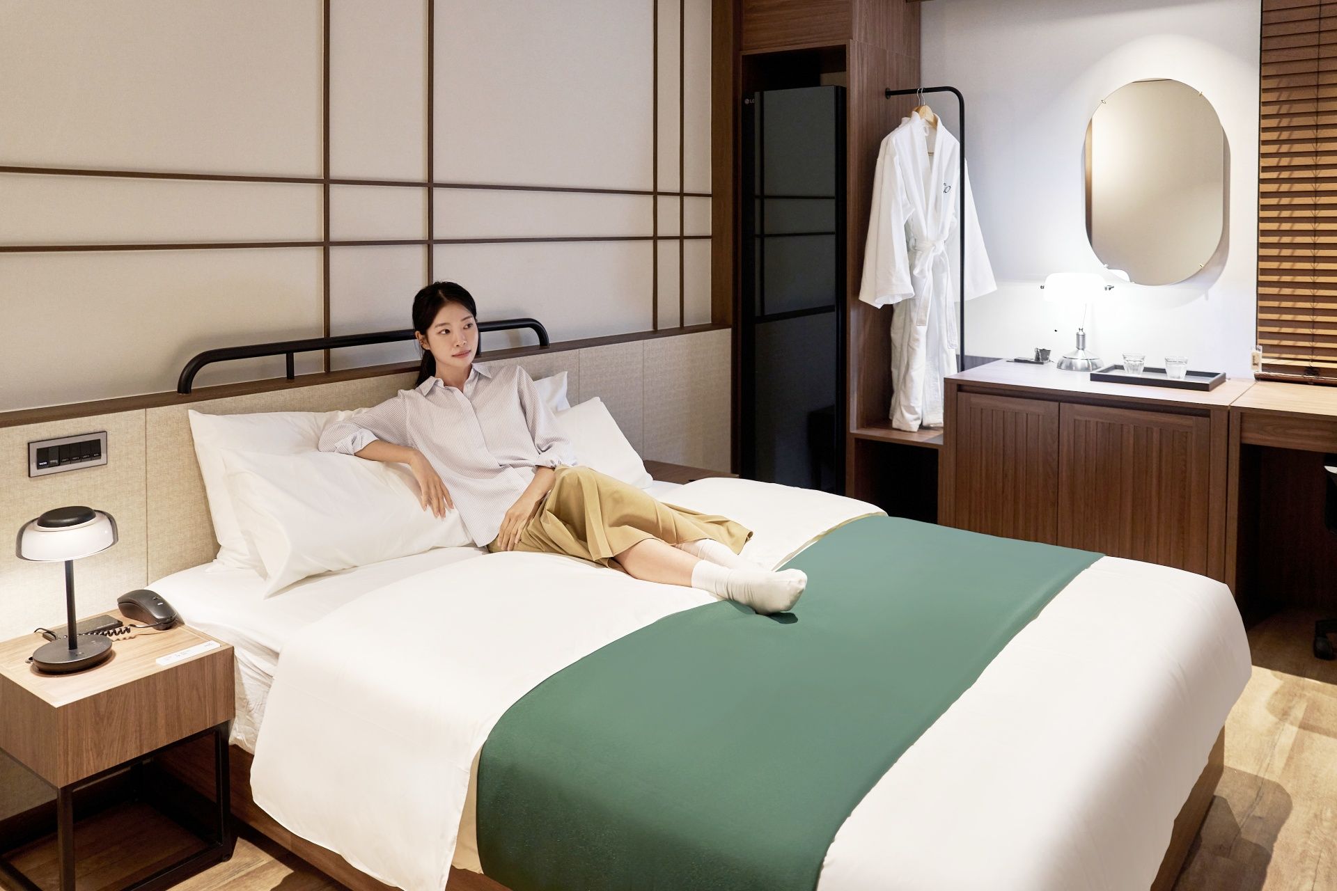 A hotel room with a bed, a bedside table with a lamp and phone, a mirror, and a wooden dresser with a robe and toiletries.