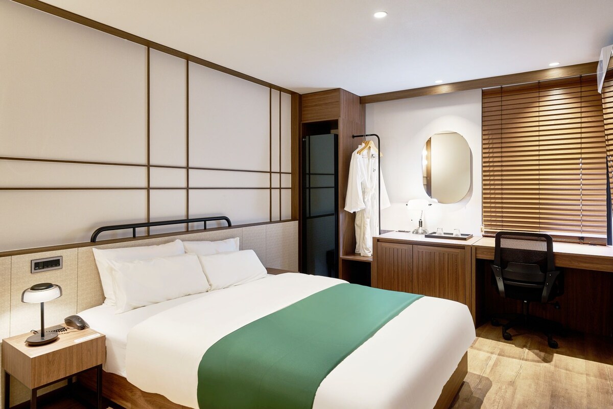 Habitación de hotel con cama, mesita de noche, escritorio, silla, espejo y ventana con persianas.